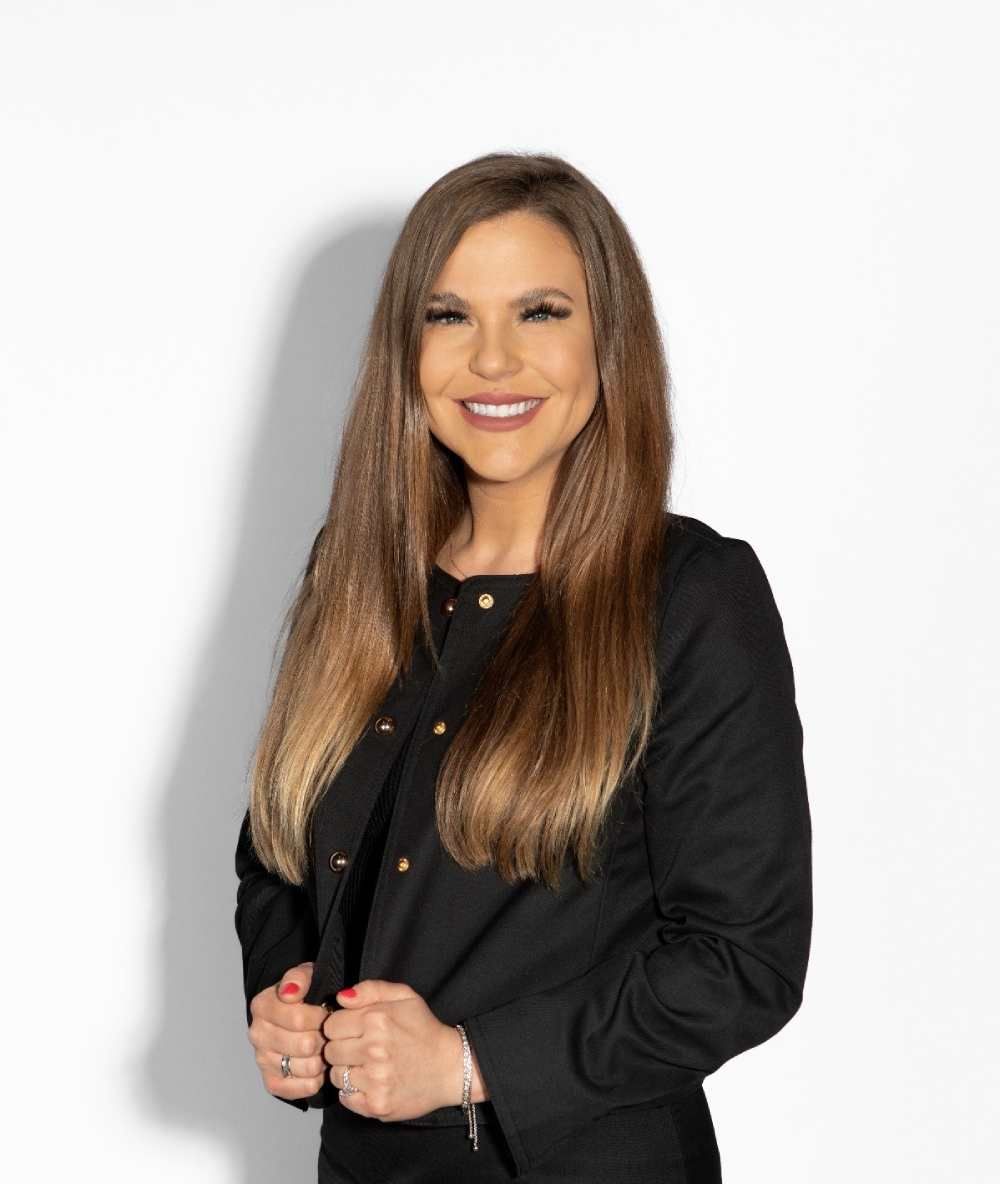 Woman with long brown hair wearing a black jacket, smiling. Against a white backdrop.