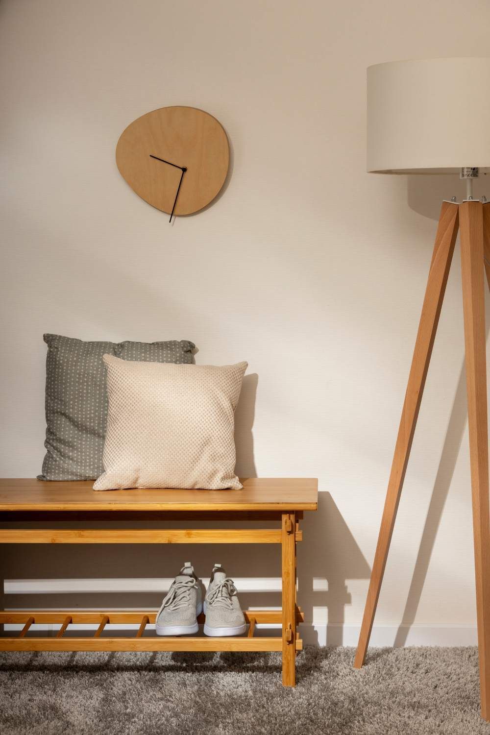 Wooden bench with pillows, clock, shoes, and a floor lamp against a white wall.