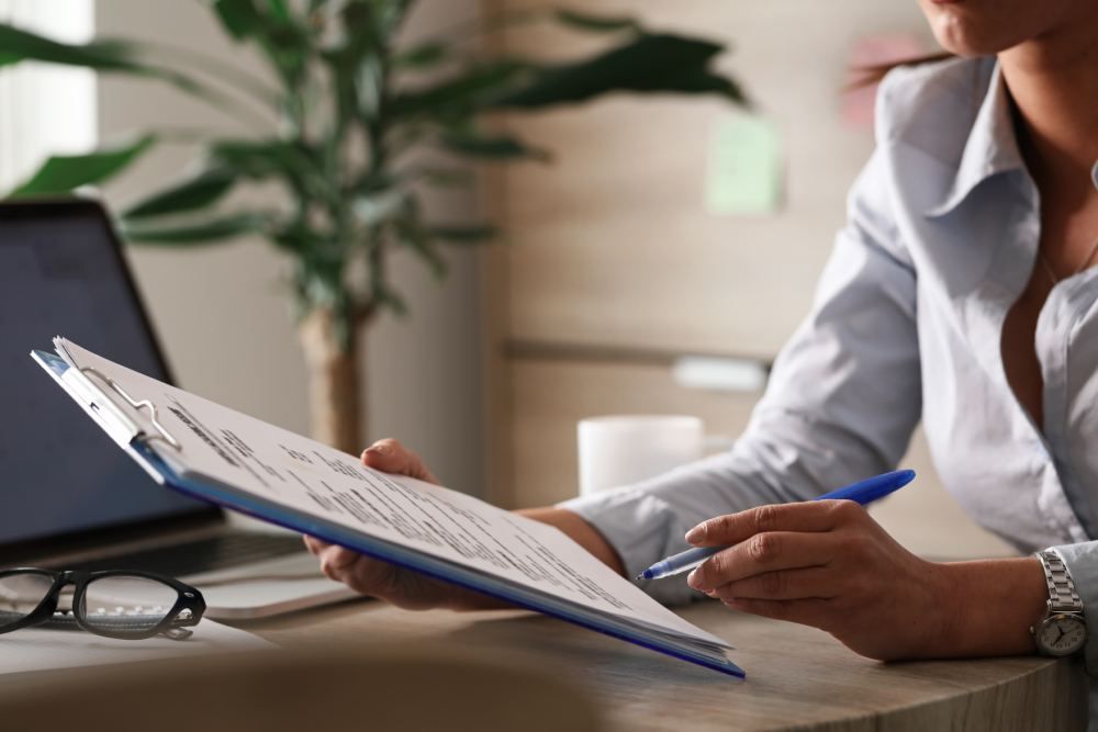Woman in blue shirt reviewing documents on clipboard at a desk. Laptop, glasses, and plant visible.