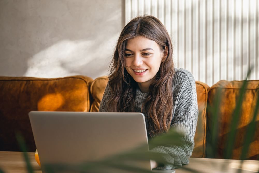 Woman smiling at laptop on table, sitting in front of an orange couch. Sunlit room.