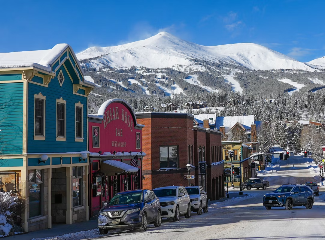 Snowy street with colorful buildings and a snow-covered mountain in the background. Cars parked along the street.