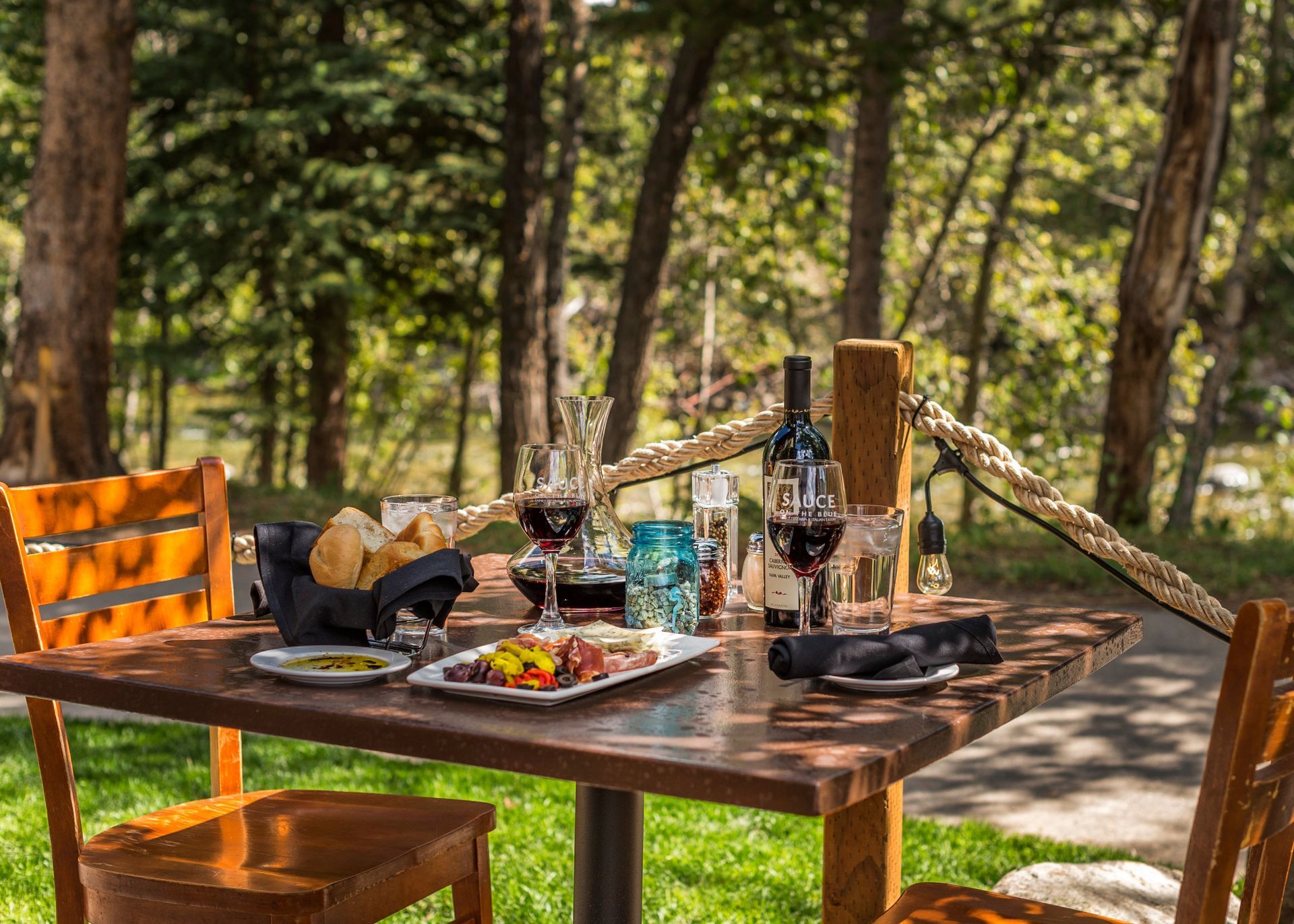 Outdoor dining table with food, wine glasses, and a wine bottle in a forest setting.