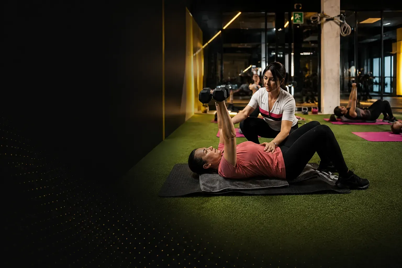 Dos personas haciendo ejercicio sobre colchonetas en un gimnasio; una ayuda a la otra a levantar pesas.