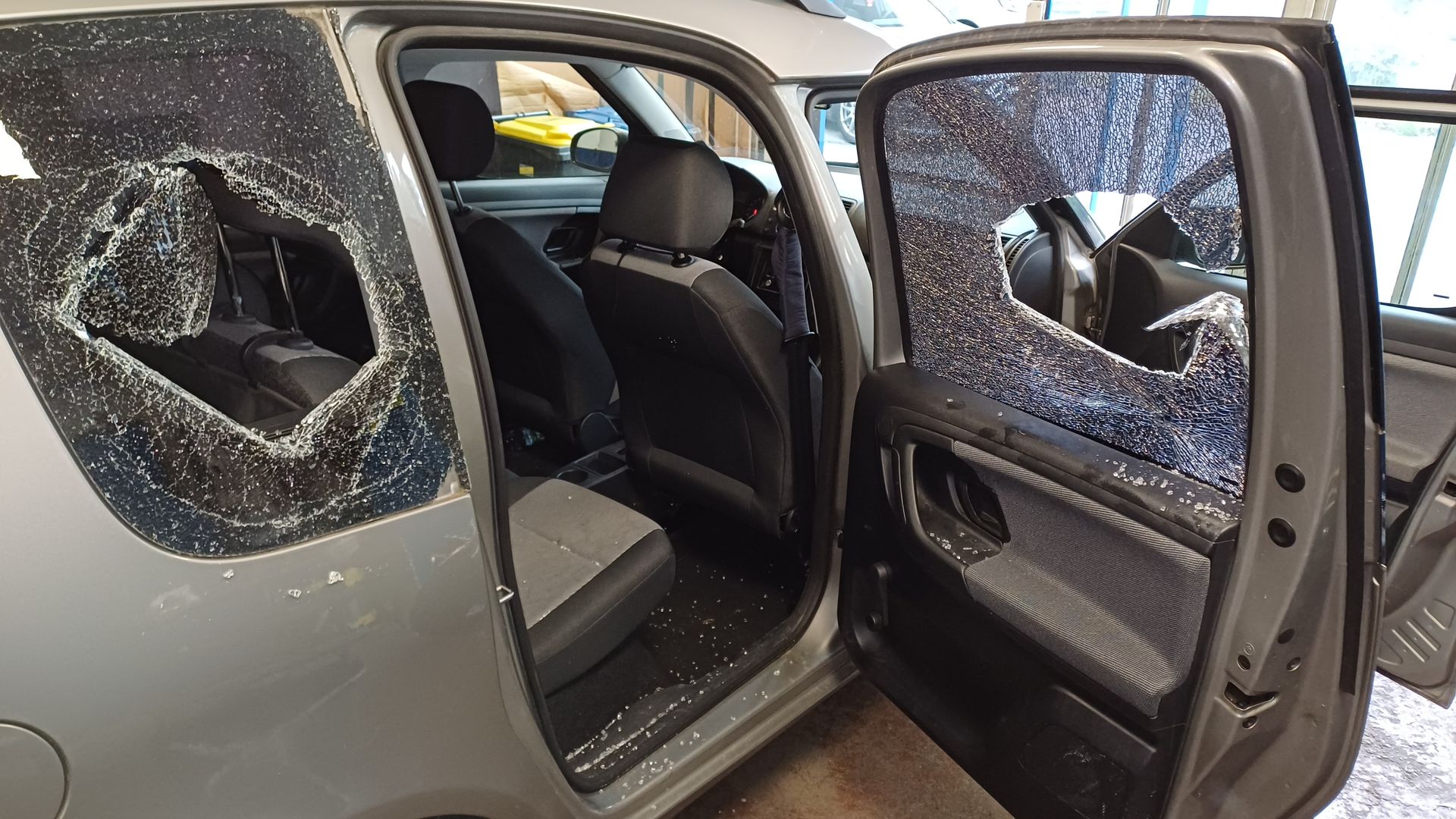 Silbernes Auto mit zerbrochenen Seitenscheiben und offenen Türen, sichtbare Glassplitter und beschädigter Innenraum