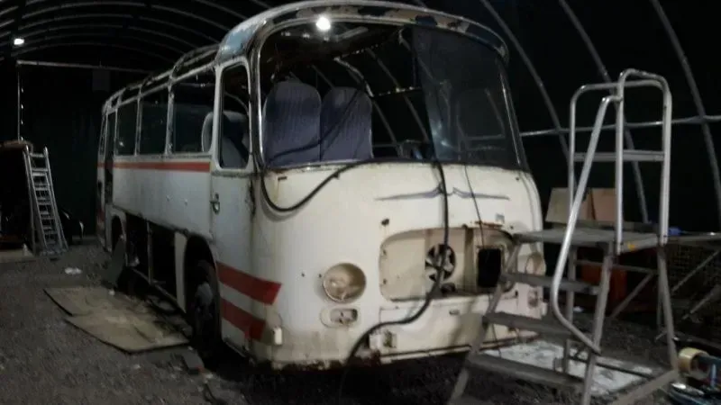 Alter weißer Bus mit roten Streifen, nachts auf einem dunklen Parkplatz abgestellt, die Front beschädigt und Teile fehlen