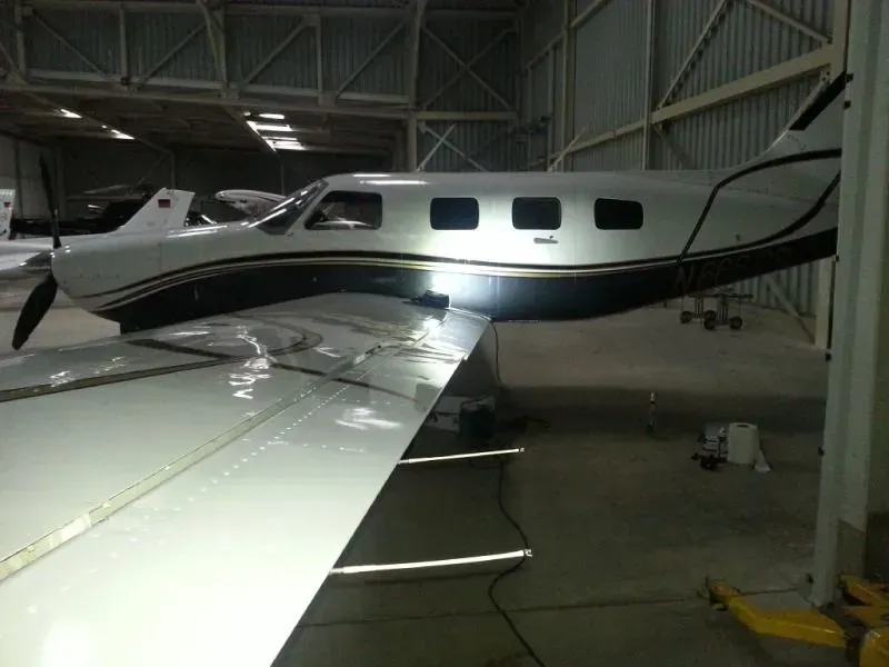 Kleines weißes Flugzeug mit dunklem Streifen, das in einem Hangar geparkt ist, aus der Perspektive des Flügels betrachtet.