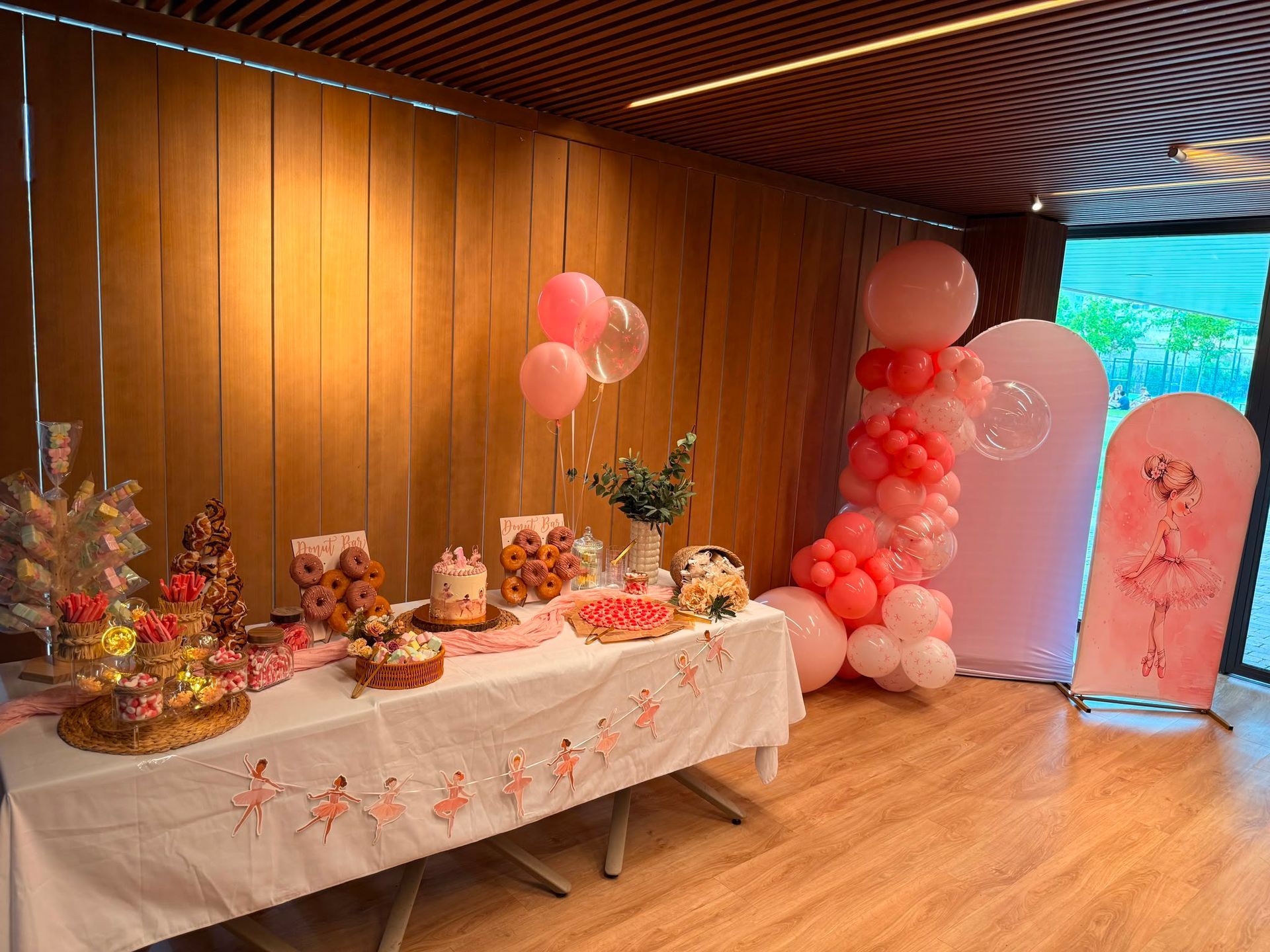 Mesa de postres de cumpleaños con globos rosas y un fondo decorativo en una habitación con paneles de madera.