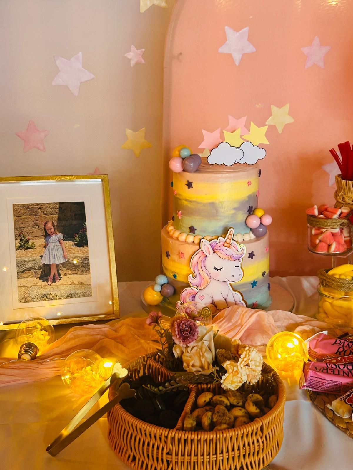 Mesa de postres de cumpleaños decorada con pasteles en tonos pastel, luces de hadas, una foto enmarcada 