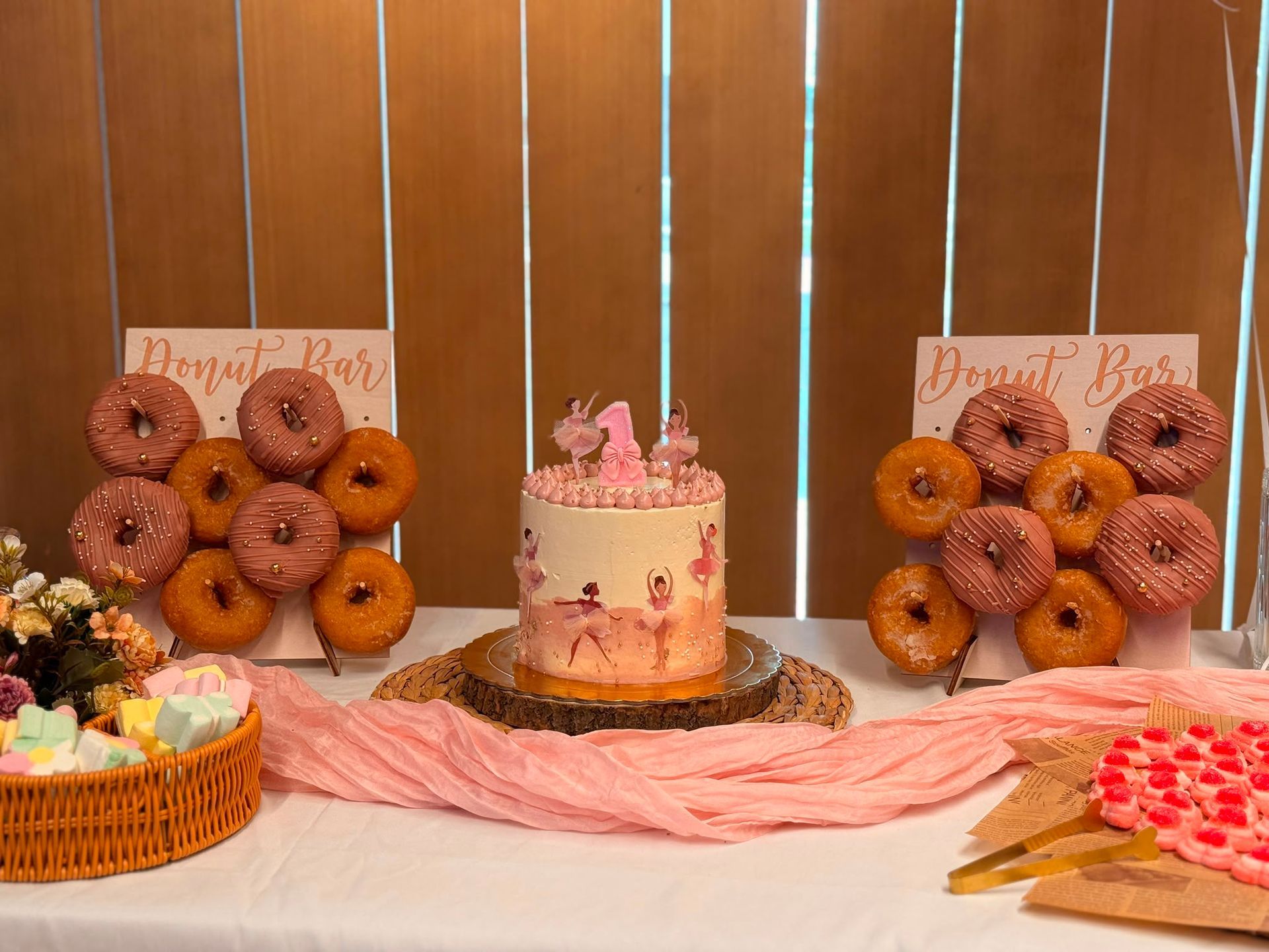 Exhibición de postres rosas con pastel, donas y cupcakes sobre una mesa decorada con fondo de madera.