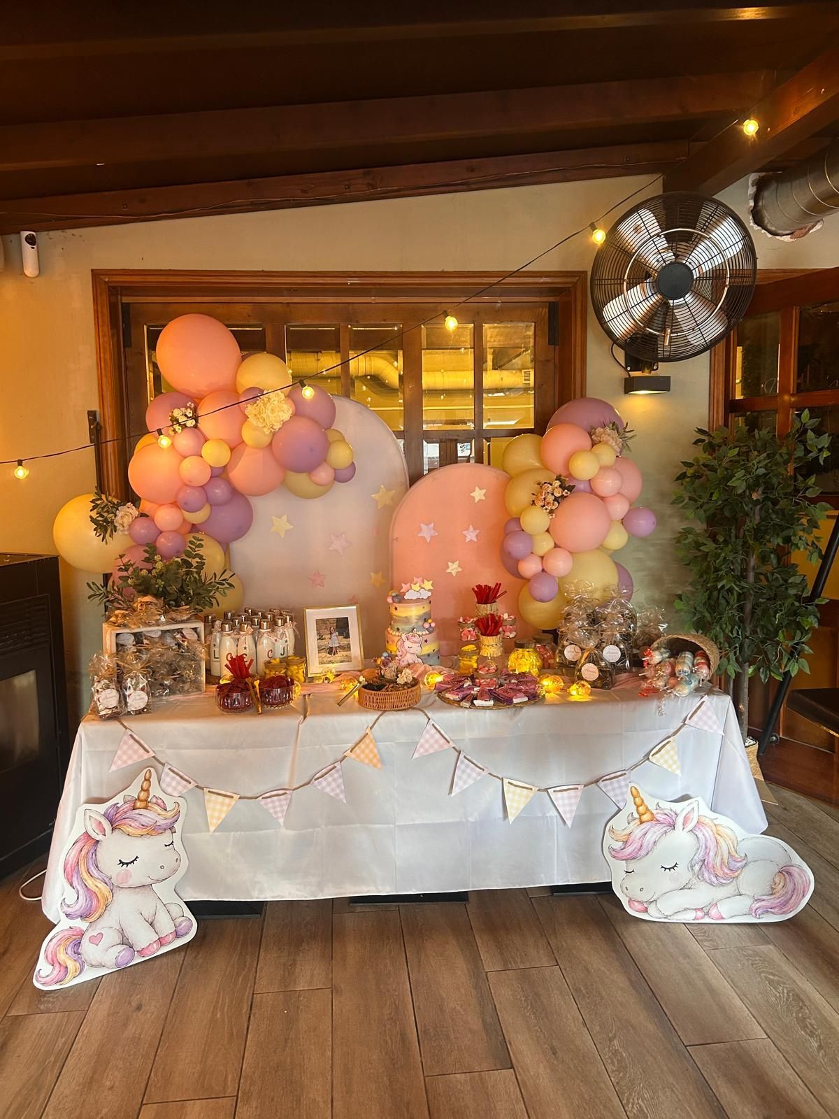 Mesa de fiesta decorada con globos en tonos pastel y figuras de unicornios en un ambiente cálido de interior.