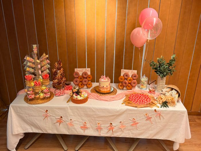 Mesa de postres con decoraciones rosas, globos, pasteles, galletas y un mantel blanco contra una pared revestida de madera.