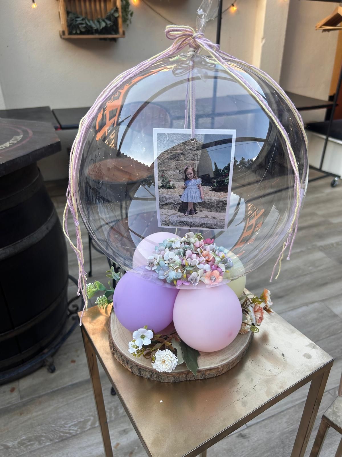 Exhibición transparente de globos con globos de color lavanda y rosa, flores y una foto enmarcada sobre una mesa pequeña.