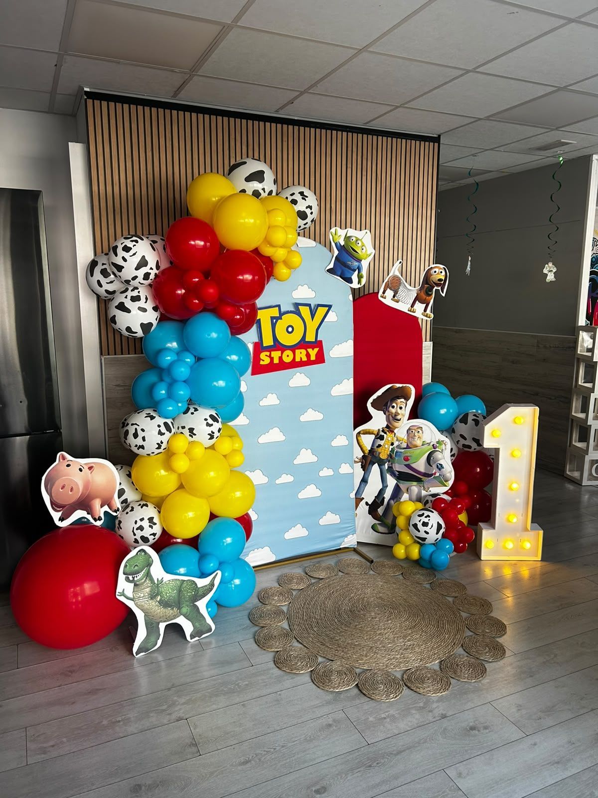 Exhibición de globos con temática de Toy Story, con globos coloridos, recortes de personajes y un letrero luminoso en un telón de fondo de escenario.