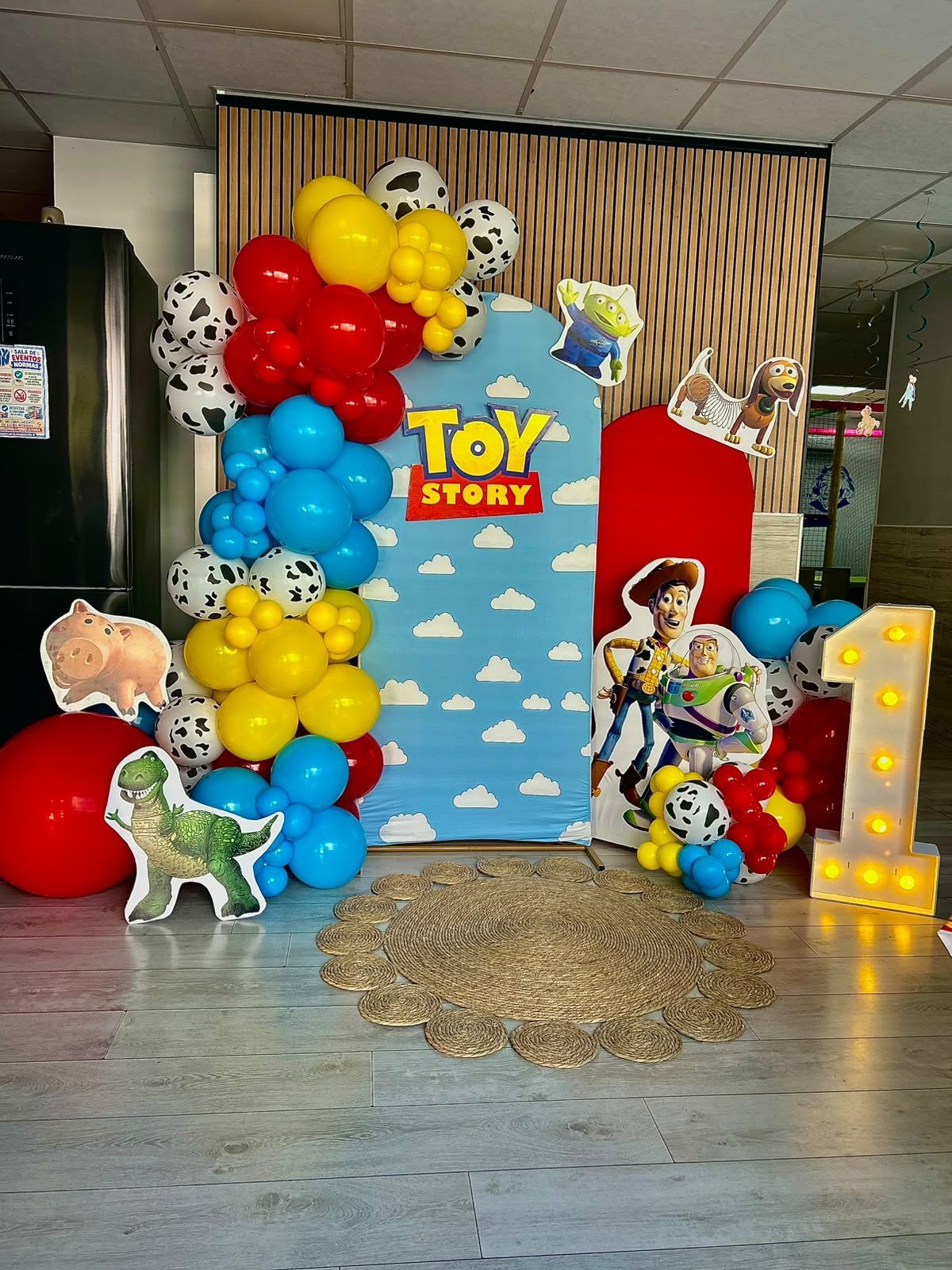 Fondo para fiesta con temática de Toy Story con globos, recortes de personajes y números luminosos tipo marquesina sobre un suelo gris.