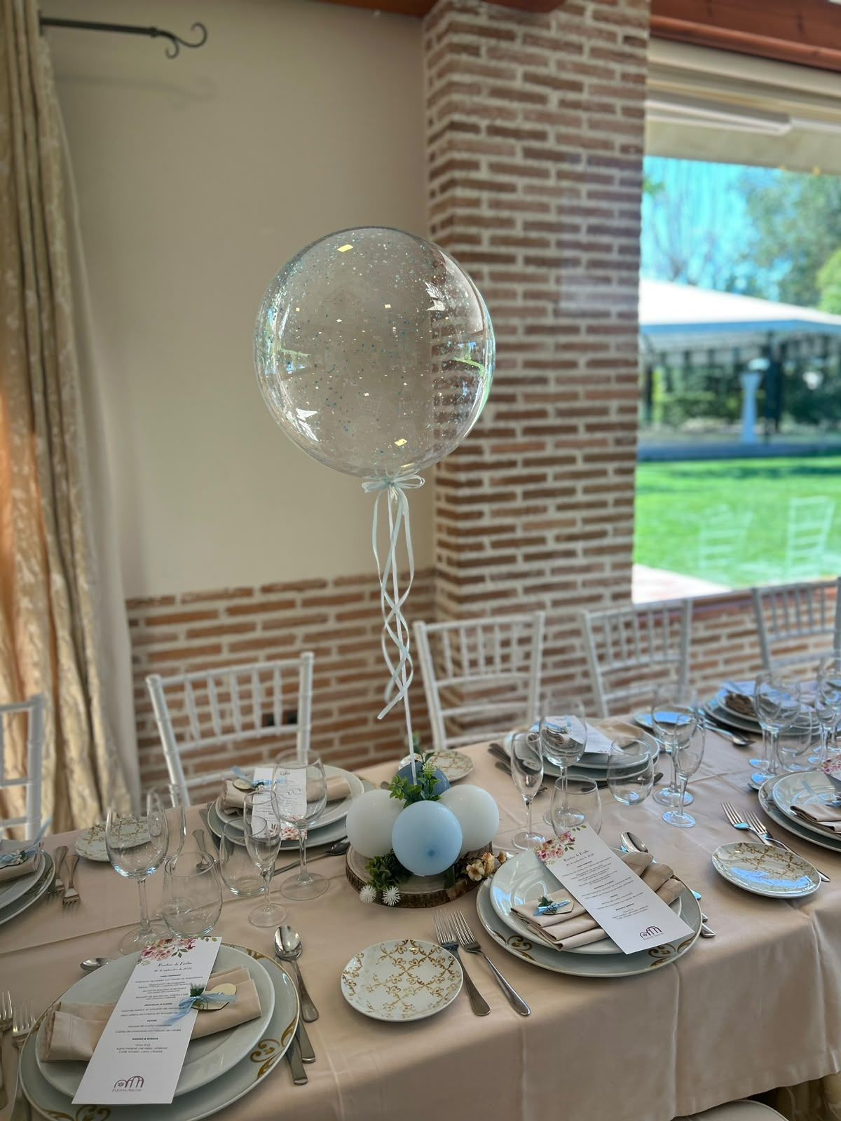 Elegante mesa de banquete con decoración en tonos beige, globos blancos, platos y vegetación junto a una gran ventana.