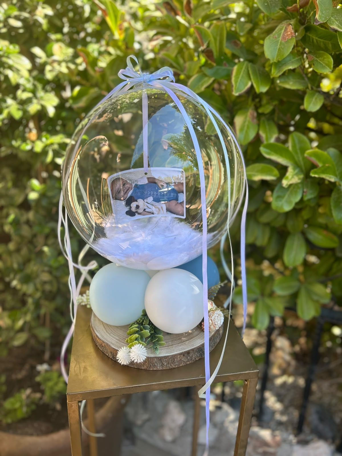 Exhibición de huevos de Pascua en una jaula colgante transparente con huevos de colores pastel y una pequeña foto, colocada sobre un taburete al aire libre.