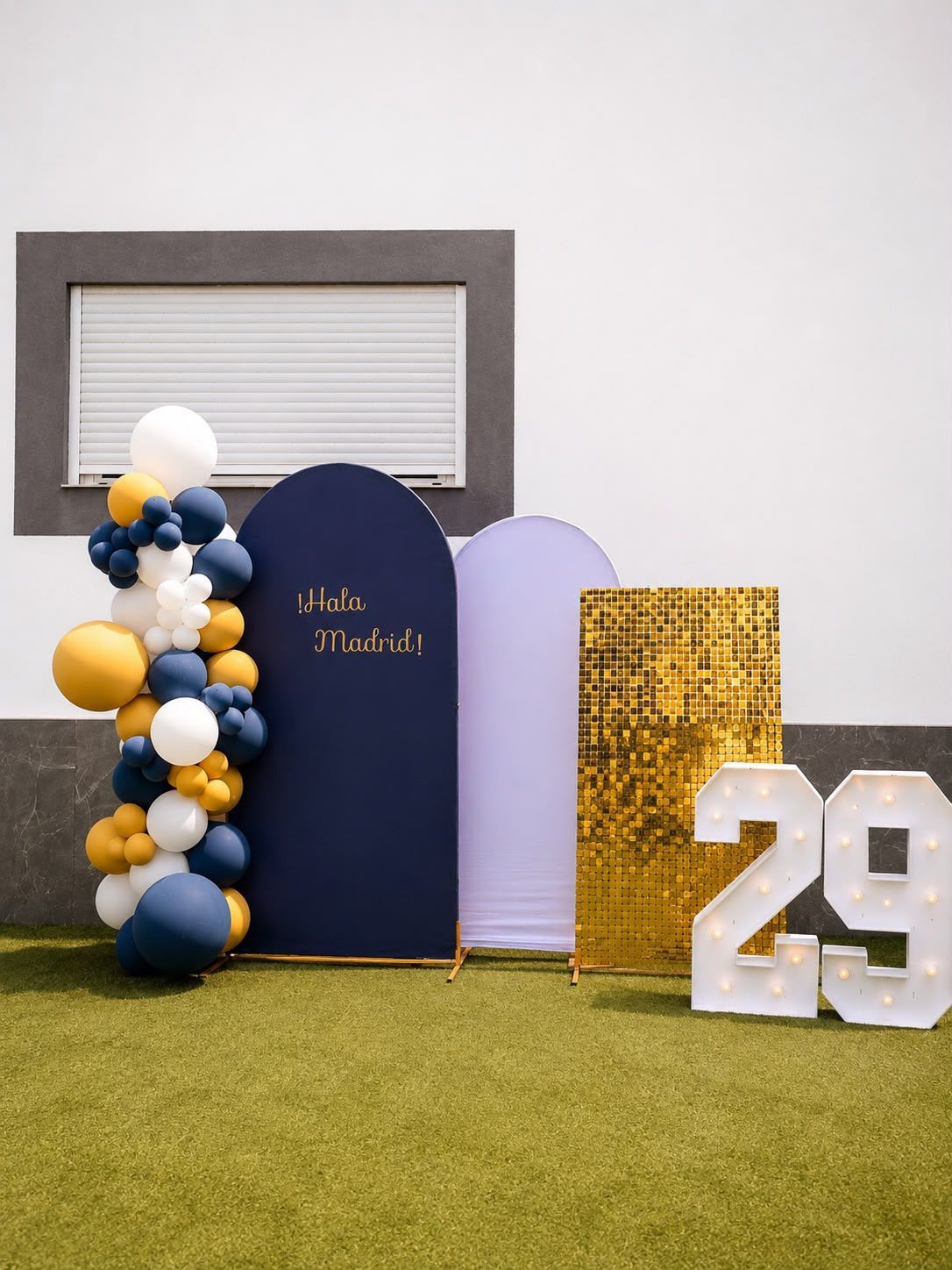 Fondo para fiesta de cumpleaños con arcos azul marino, lavanda y dorado, guirnalda de globos y un “29” blanco sobre césped.