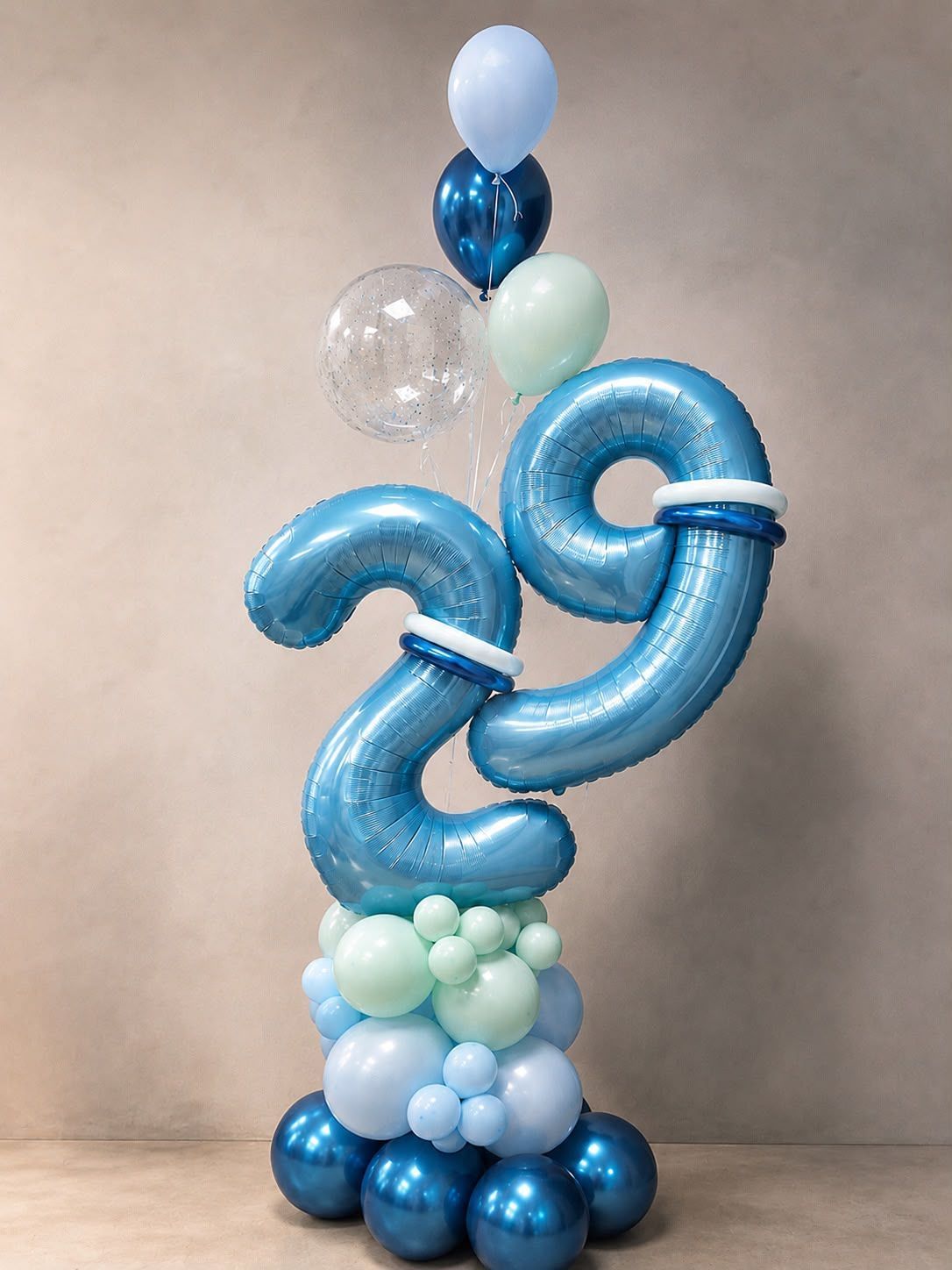 Escultura de globos azules con grandes números entrelazados, adornada con globos azul claro, blancos y transparentes sobre un pedestal.