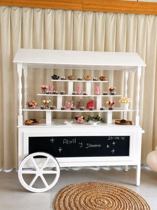 Un carrito de postres de madera blanca con estantes llenos de cupcakes y flores, situado frente a unas cortinas color crema