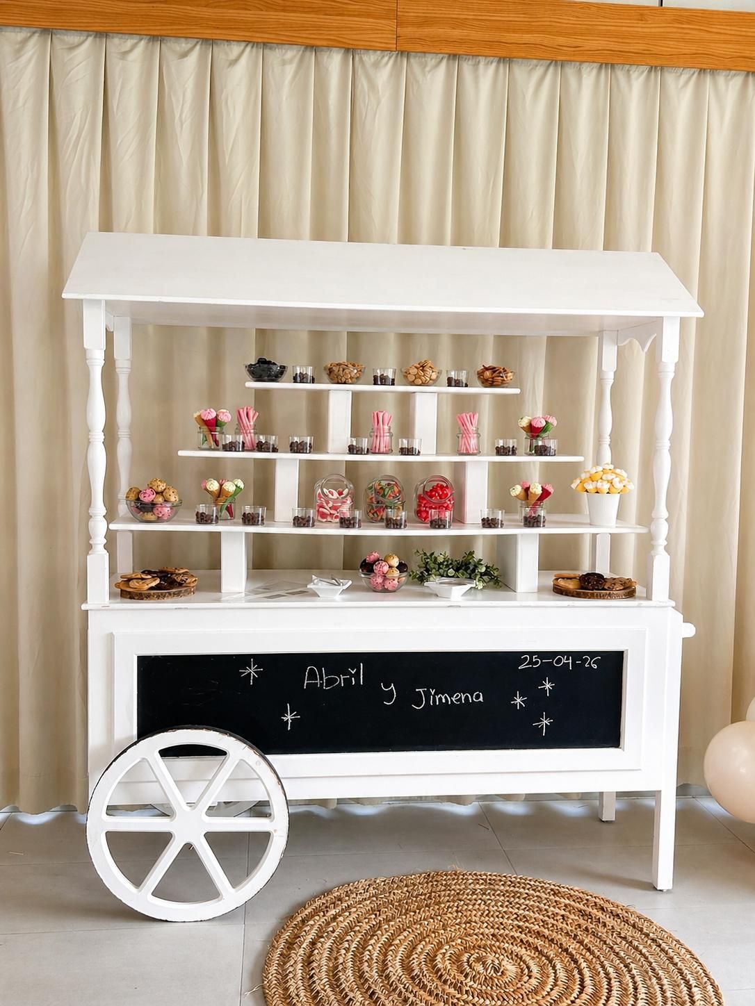Carrito de postres blanco con estantes repletos de dulces florales y una pizarra en la parte frontal