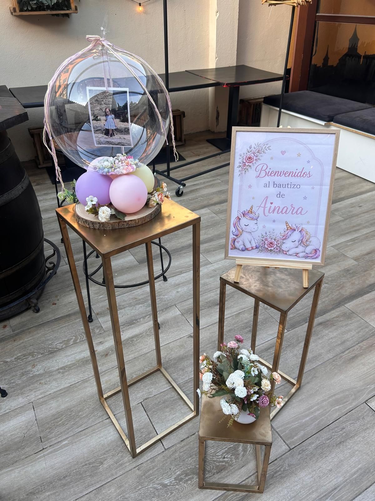 Exhibición exterior decorada con un globo transparente, flores y un letrero con marco rosa sobre soportes dorados.