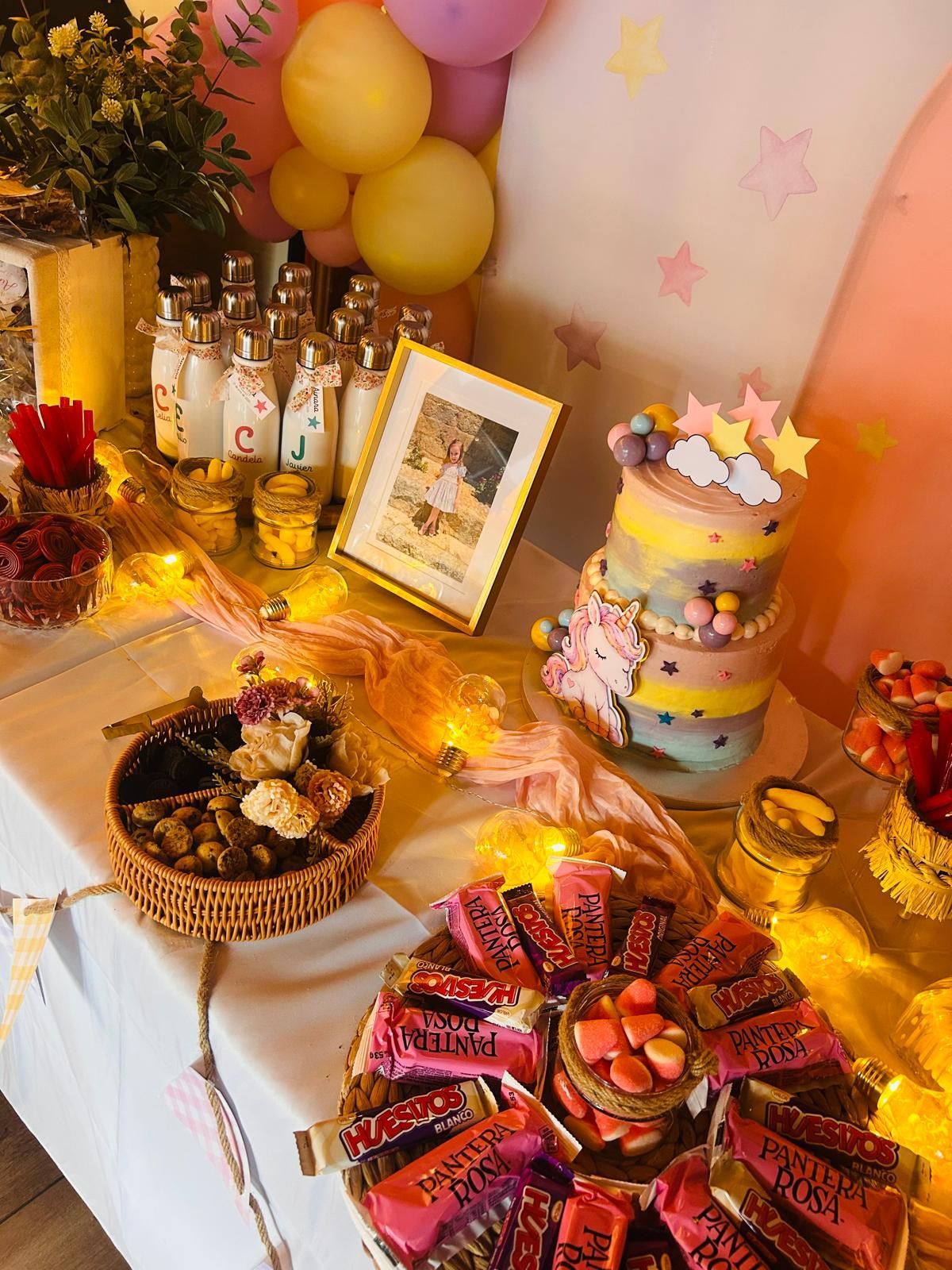 Mesa de postres de cumpleaños con globos, pastel, dulces y luces de hadas cálidas.