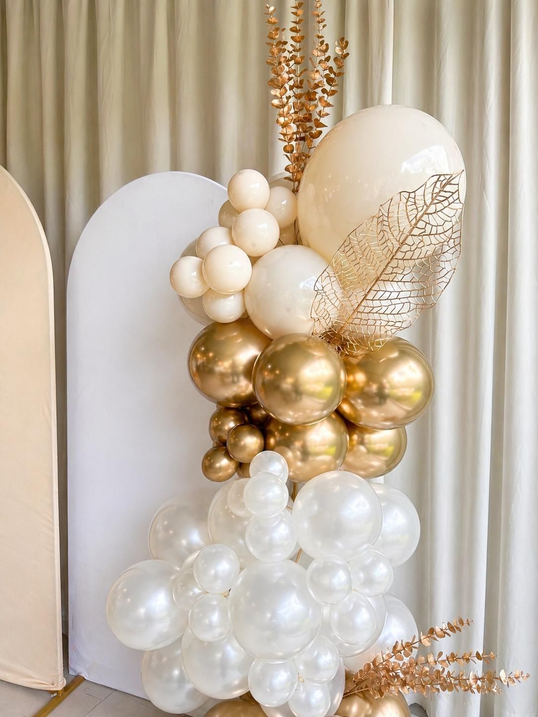 Arreglo de globos en tonos crema, blanco y dorado con detalles florales secos sobre un fondo blanco.