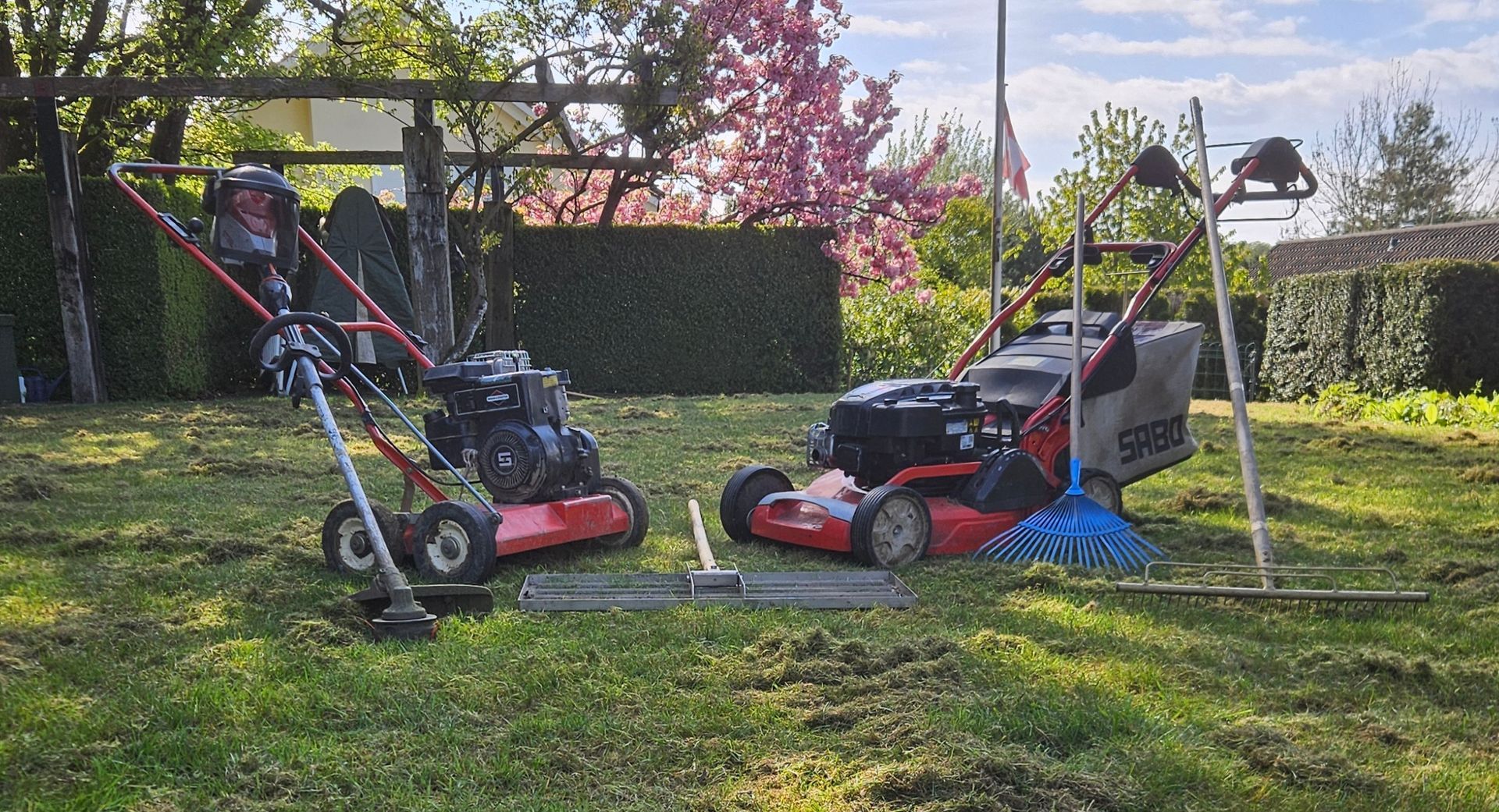 Zwei Rasenmäher auf einer sonnigen Rasenfläche, einer in der Nähe eines Metallrahmens und der andere bei einer Hecke.