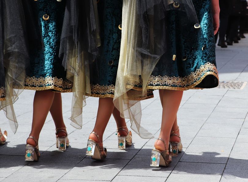 Piernas de mujeres con faldas verde azulado y tacones con detalles dorados caminando sobre adoquines.