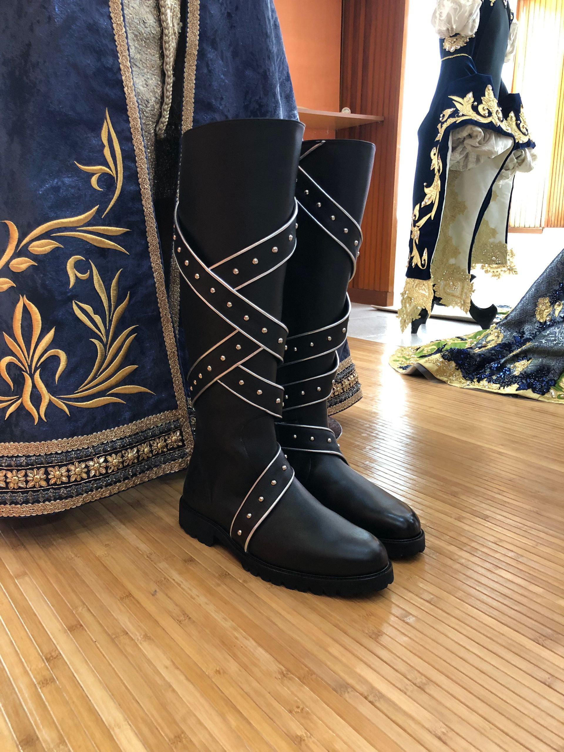 Botas negras hasta la rodilla con tiras tachonadas de plata sobre un piso de madera, con ropa azul adornada visible.