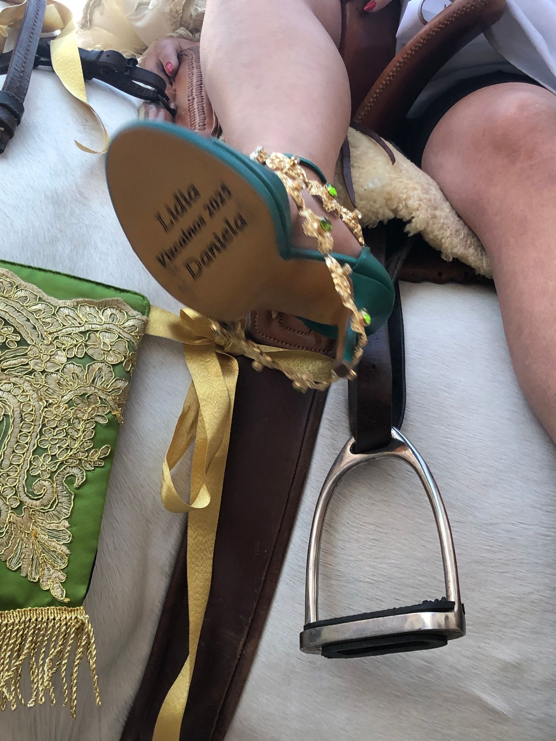 Un pie con un zapato verde con detalles dorados reposa en un estribo. La suela lleva grabado