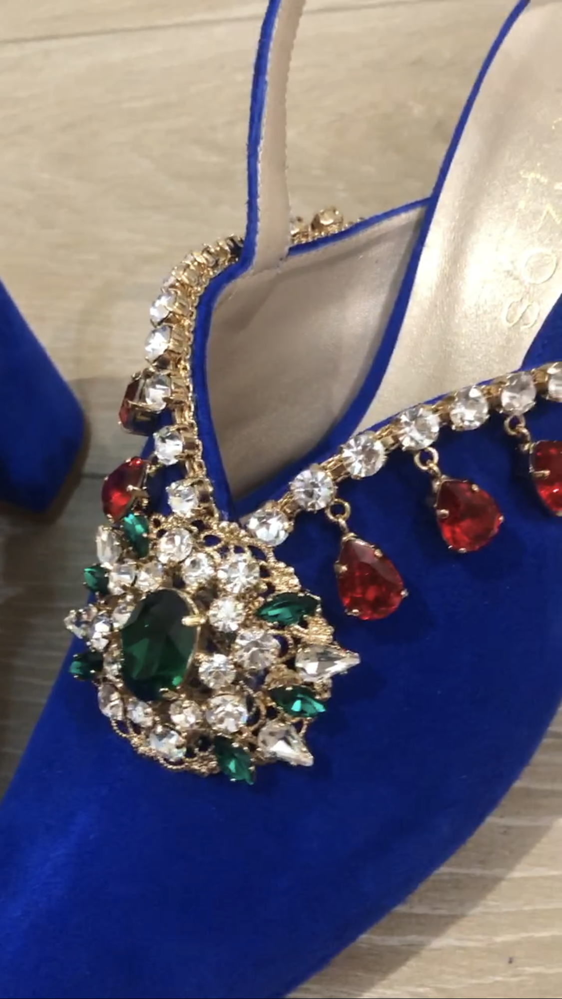 Zapato de tacón alto con joya azul, con una gran gema verde y blanca y joyas rojas en forma de lágrima.