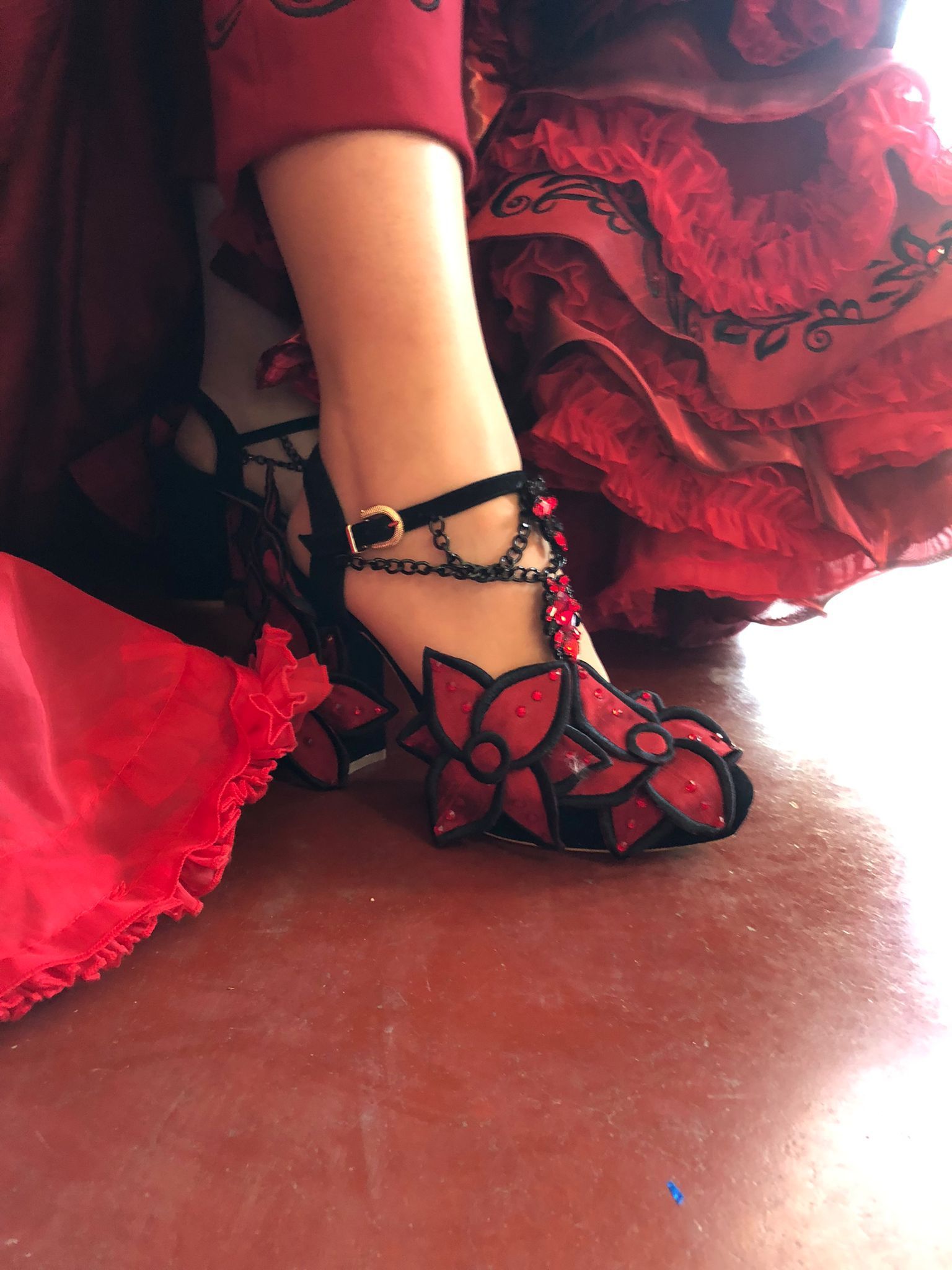 Zapato de flamenco rojo con diseño floral, detalle de cadena negra, sobre suelo de baldosa roja.