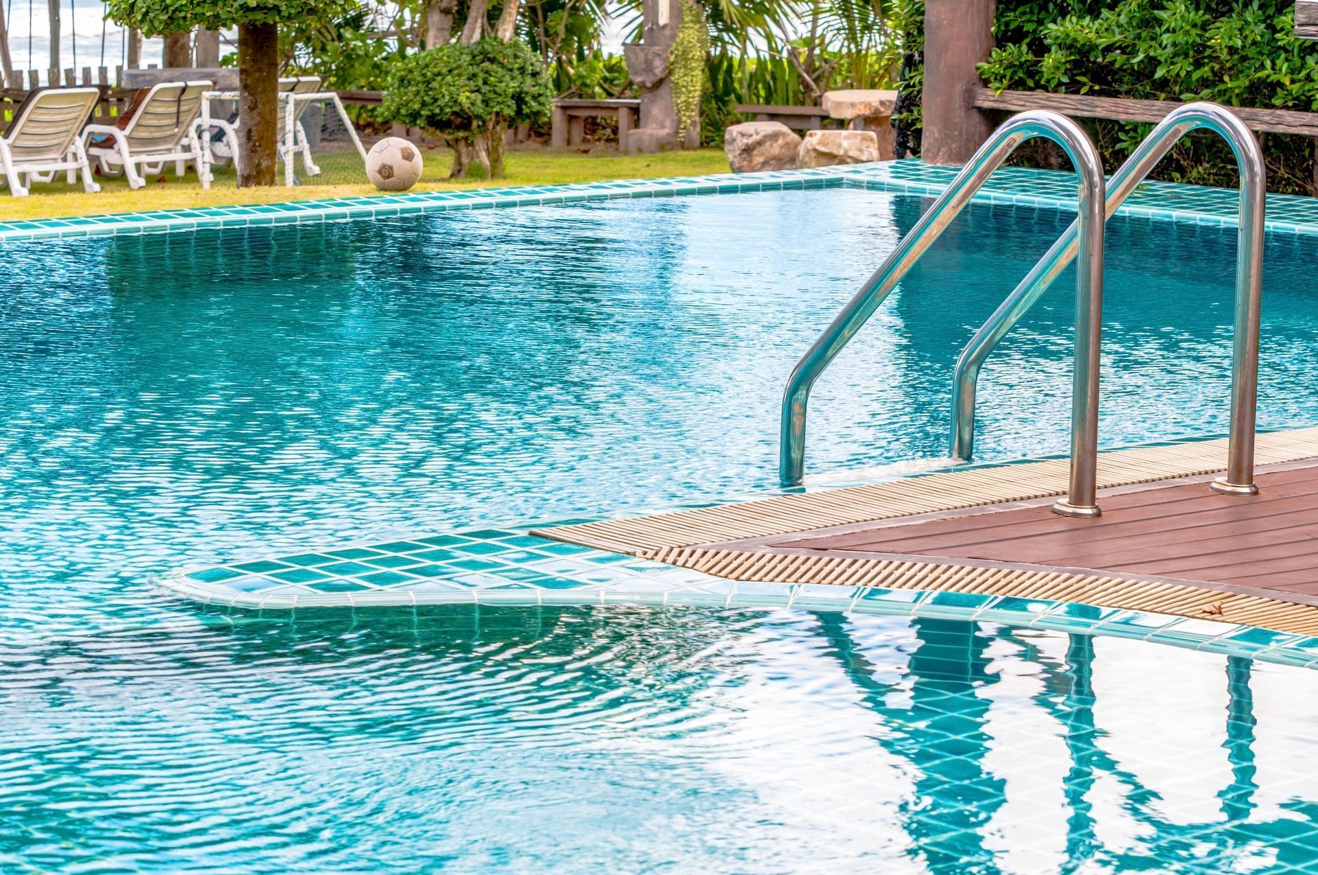 Piscina con escalera cromada, agua turquesa, borde de ladrillo y césped al fondo.