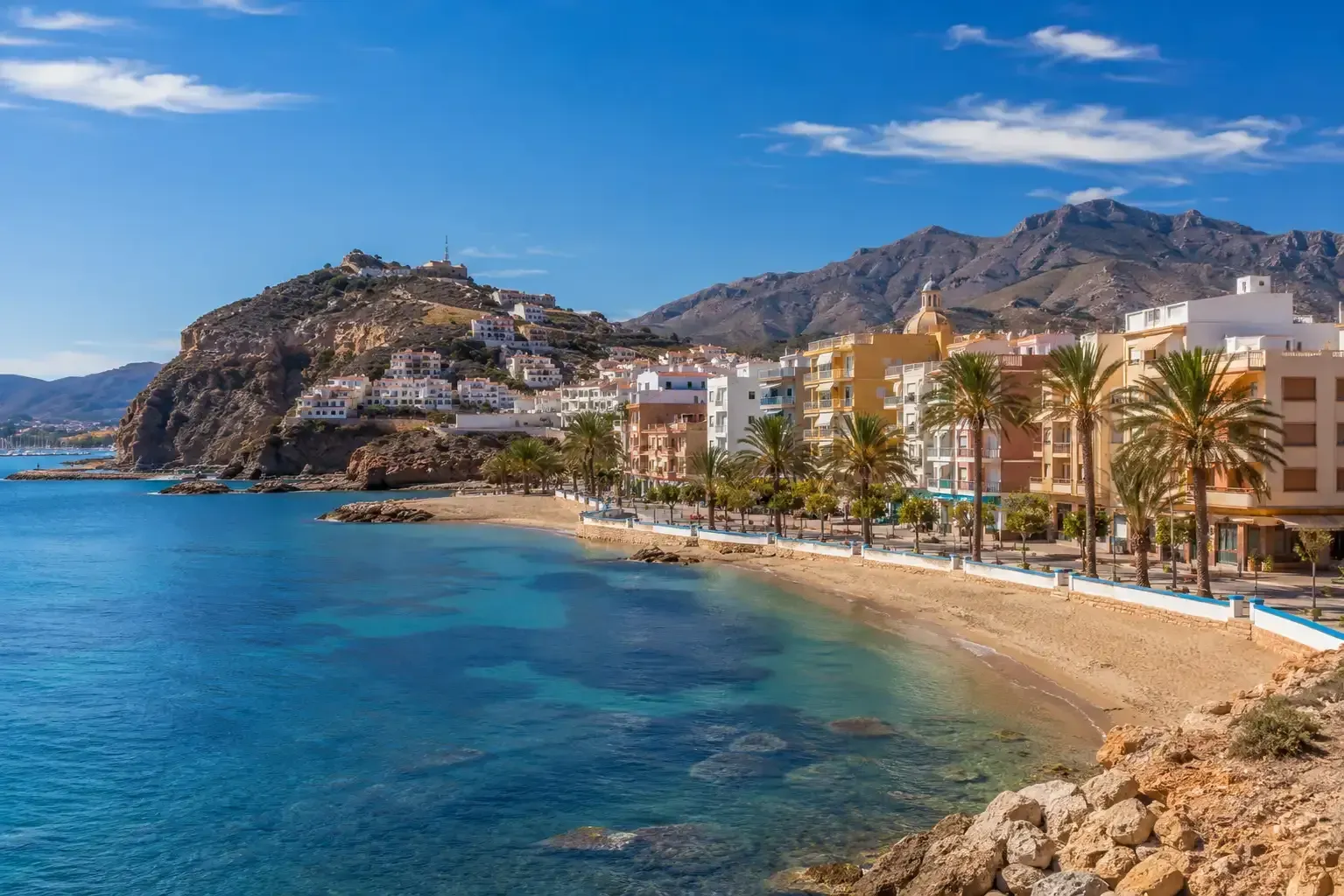 Bahía costera soleada con aguas turquesas, playa de arena, palmeras y edificios en la ladera bajo un cielo despejado.