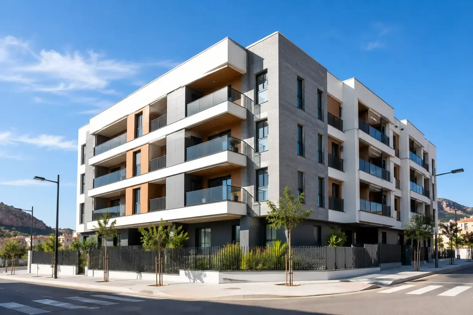 Edificio de apartamentos moderno en una esquina soleada, con balcones y acera ajardinada.