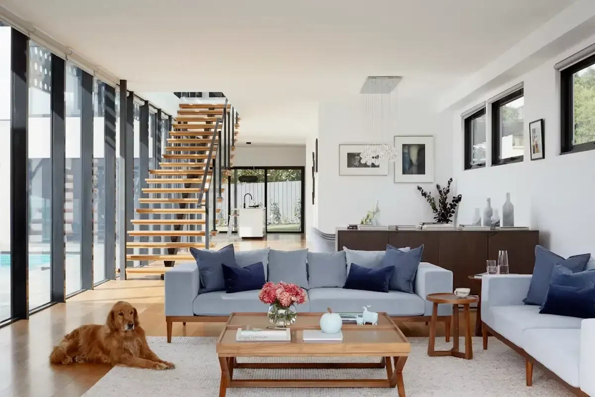 Luminoso salón moderno con sofás azules, escalera de madera y un perro tumbado en el suelo.