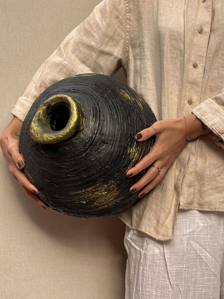 Una persona vestida con un atuendo de lino beige sostiene una vasija redonda de cerámica negra texturizada con detalles pintados en dorado.