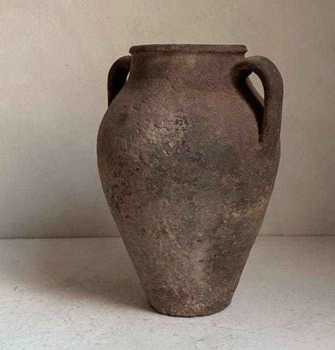 Un jarrón rústico de arcilla marrón con dos asas y una superficie texturizada y desgastada, centrado sobre un fondo liso de color claro.