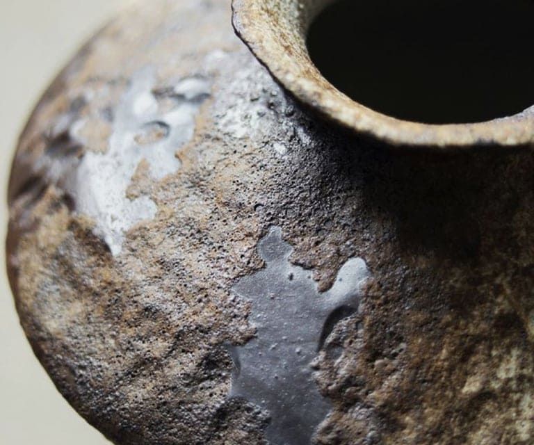 Primer plano de una vasija de cerámica rústica y texturizada, con manchas de esmalte oscuro y liso que contrastan con una superficie marrón rugosa.