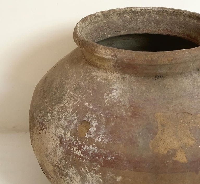 Una maceta redonda de terracota, desgastada por el tiempo y con un borde ancho, que presenta pátinas de color marrón tierra, rojo apagado y blanco texturizado.