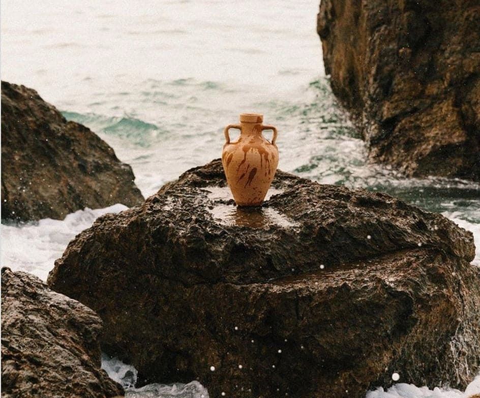 Un jarrón de cerámica de color tostado con dos asas descansa sobre una gran roca oscura rodeada por el oleaje del mar.