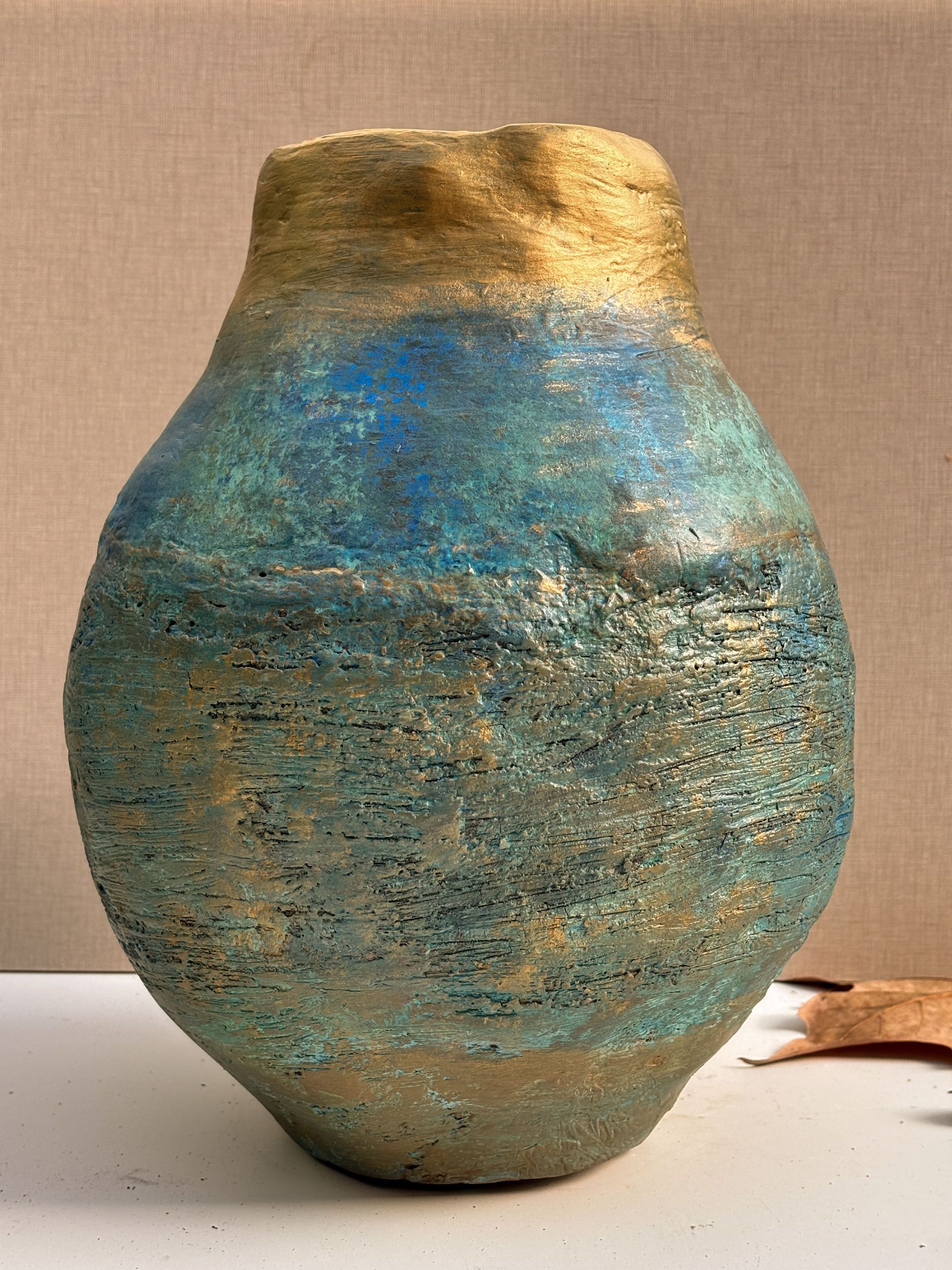 Un jarrón redondo de cerámica texturizada con borde y base de color dorado metalizado, que presenta una superficie moteada en azul y dorado.