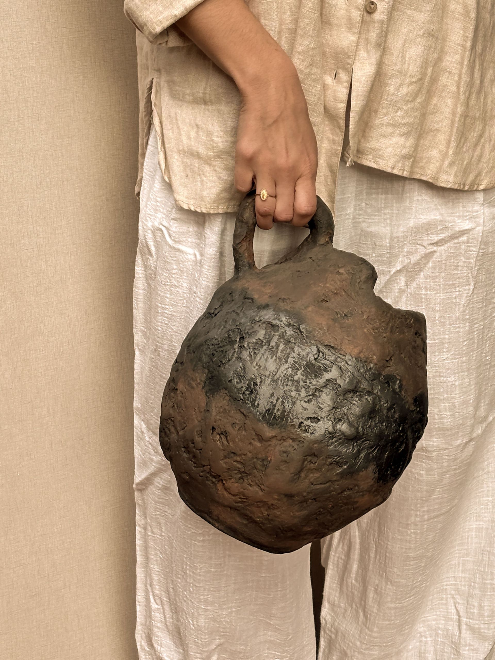 Una mano sostiene un recipiente de cerámica rústico, redondo, de color marrón oscuro y con textura, que tiene un solo asa, sobre una prenda de vestir de color claro.