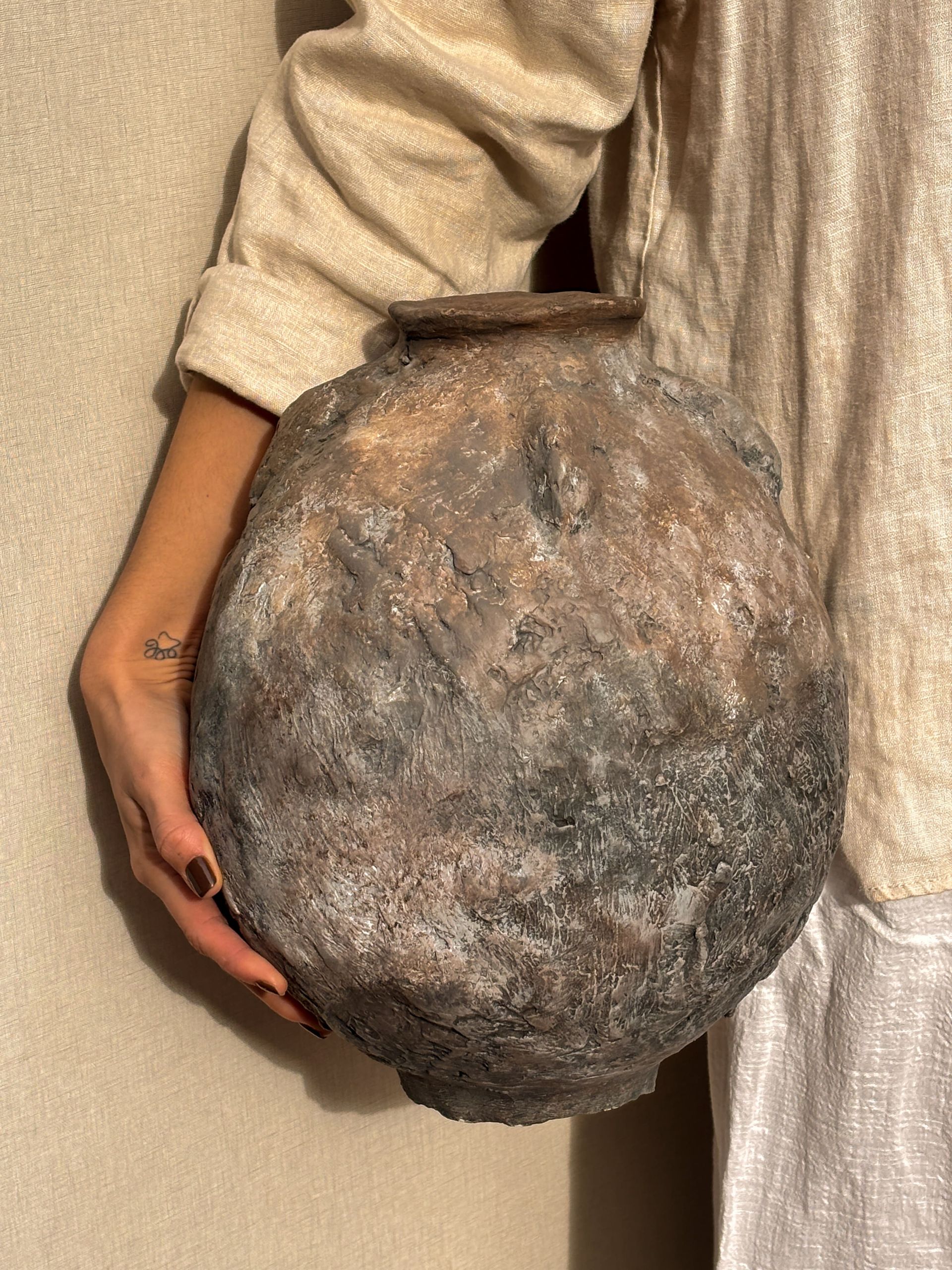 Una persona sostiene un jarrón de cerámica grande, redondo y texturizado, con un esmalte oscuro, moteado y de tonos tierra, apoyado contra una pared de color neutro.