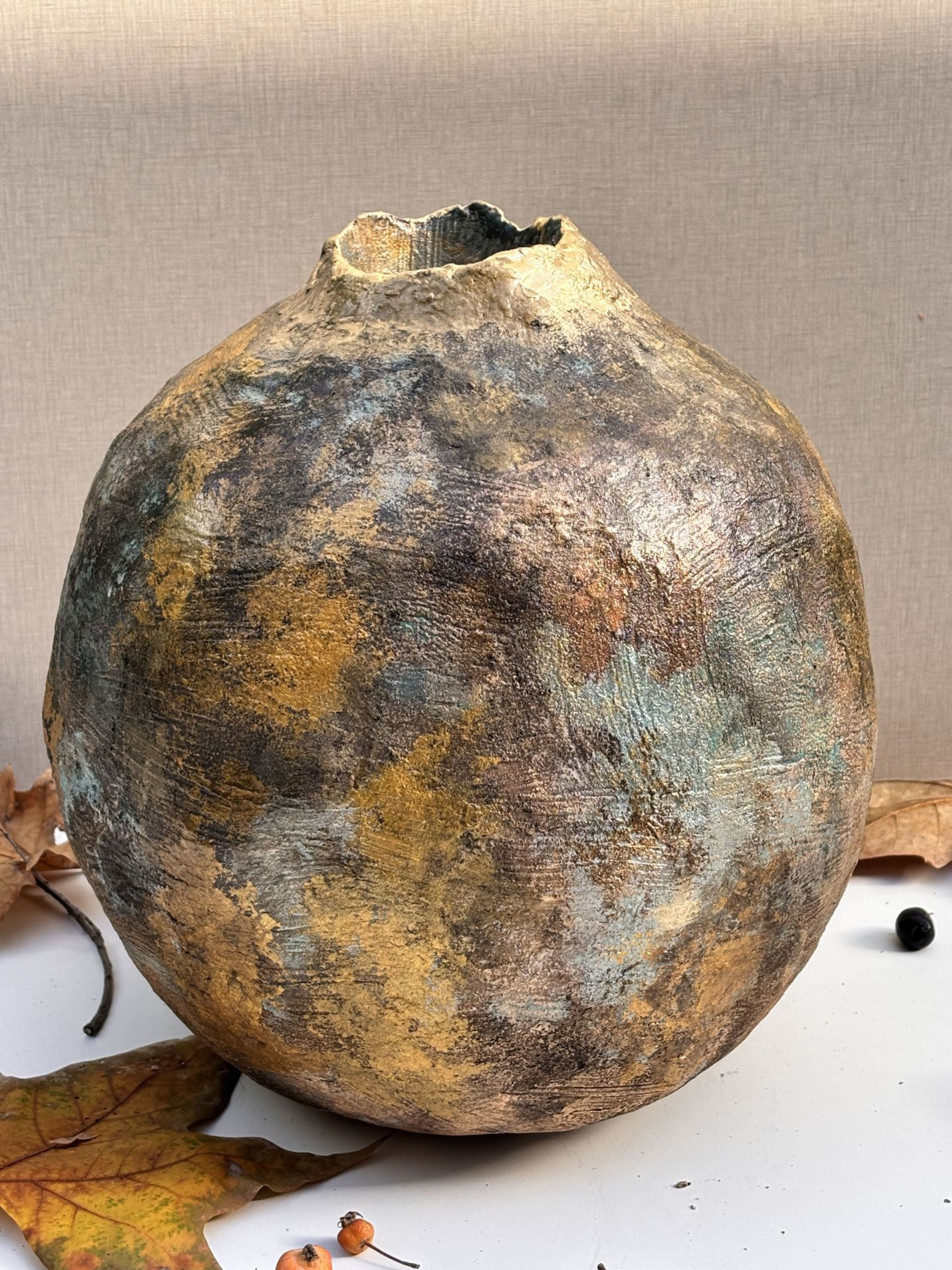 Un jarrón esférico de cerámica texturizada con un acabado moteado en oro, bronce y gris oscuro reposa sobre una superficie blanca decorada con hojas.