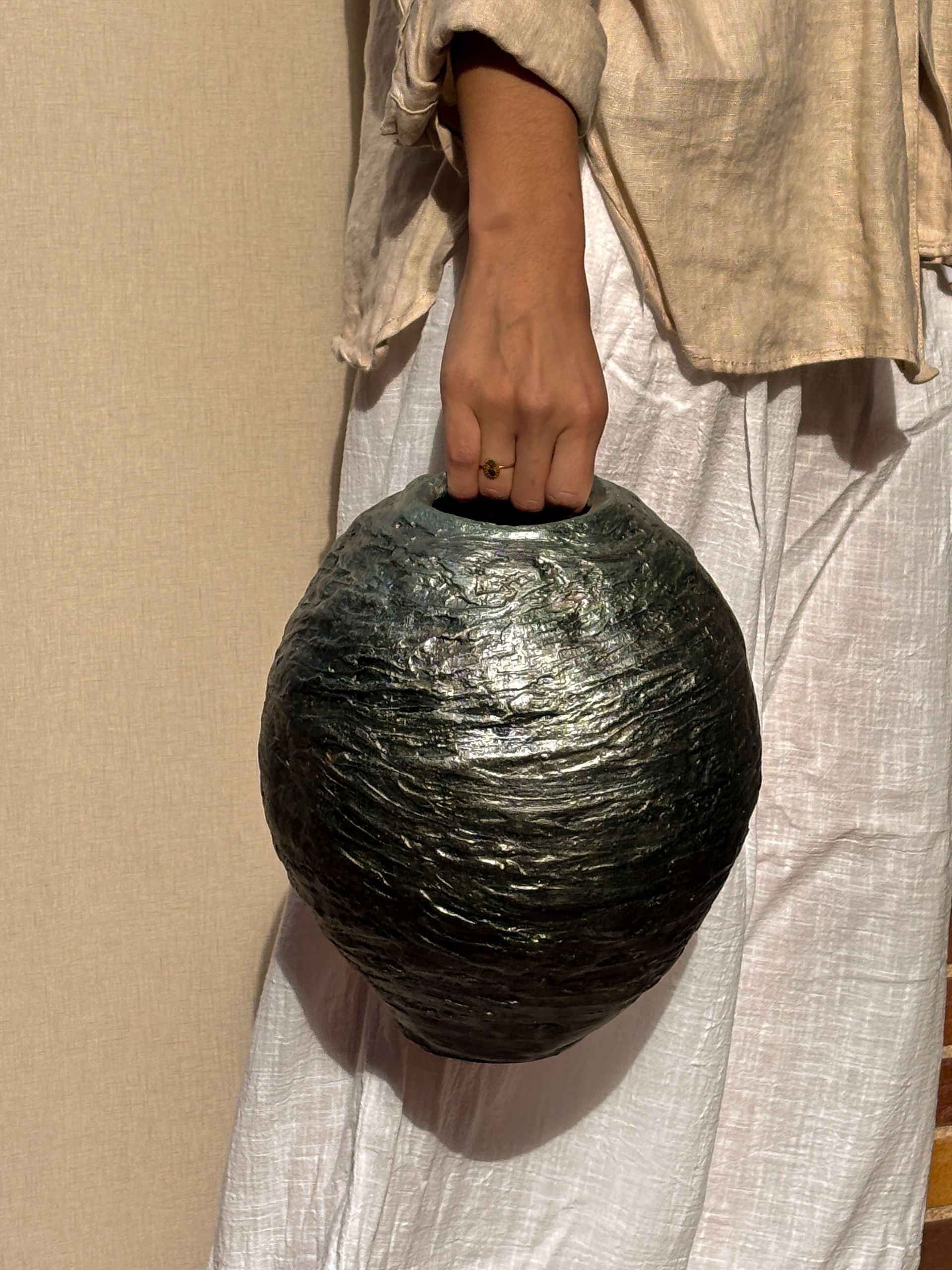 Una persona vestida con ropa de lino de color neutro sostiene un recipiente grande, esférico, texturizado y de color gris oscuro.