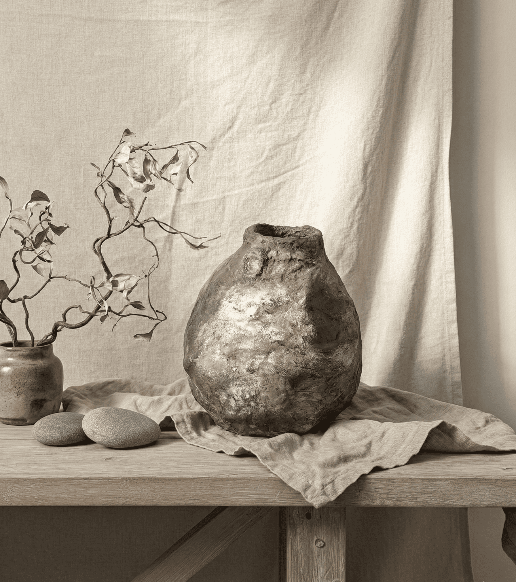 Un bodegón rústico con un jarrón de arcilla texturizada, una pequeña maceta de cerámica con ramas y dos piedras sobre una mesa de madera.