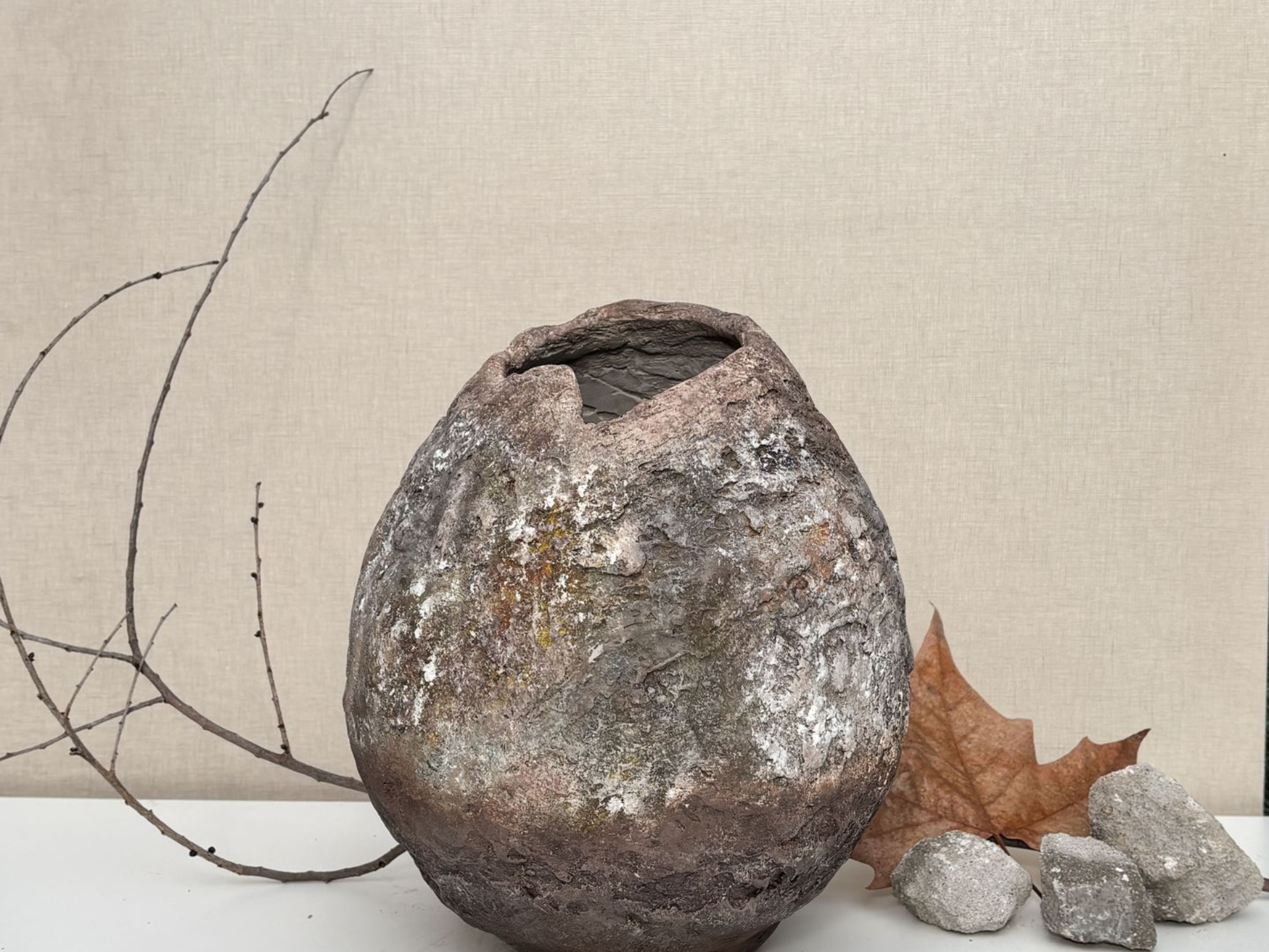 Un recipiente redondo, de arcilla oscura y con textura, de borde irregular, reposa junto a una ramita seca, una hoja marrón y tres pequeñas piedras grises.
