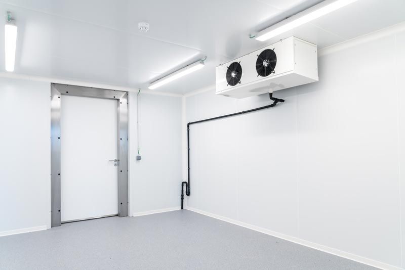 Chambre froide d'un blanc éclatant, avec une porte métallique et un groupe frigorifique mural relié par des tuyaux noirs.